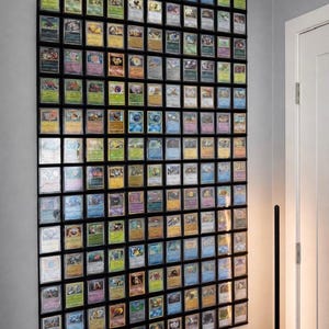 May include: A wall-mounted display of framed trading cards, arranged in a grid. The cards feature colourful illustrations and designs, set within black frames. The display is against a grey wall.