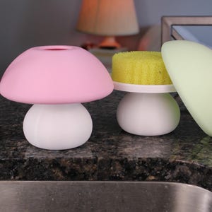 May include: Three mushroom-shaped kitchen accessories. A pink mushroom-shaped sponge holder, a white mushroom-shaped sponge holder with a yellow sponge, and a light green dish scraper. The items are on a dark speckled countertop.
