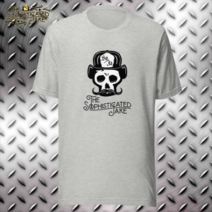 Könnte beinhalten: Hellgraues T-Shirt mit einem Totenkopf-Design, das einen Hut mit dem Text "So Ja" und einen Schnurrbart trägt. Der Text "The Sophisticated Jake" steht darunter. Das Shirt ist aus weichem Material.