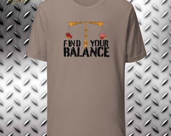 Puede incluir: Camiseta color beige con la frase "FIND YOUR BALANCE" en negro. El diseño presenta una balanza con un corazón a un lado y un cerebro al otro. La balanza es de color dorado.
