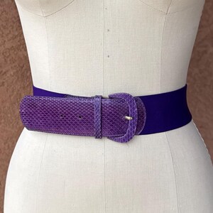 Op de afbeelding: Een paarse riem met een gestructureerde gesp met slangenleerpatroon en een gladde, elastische band. De riem is bevestigd rond de taille van een witte, torso-vormige etalagepop met de tekst COLLAPSIBLE SHOULDER en het cijfer 8 erop.