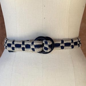 May include: A woven belt with a navy blue and beige pattern and a navy blue buckle. The belt is displayed on a mannequin with the words "COLLAPSIBLE SHOULDER" and the number "8" printed on it.