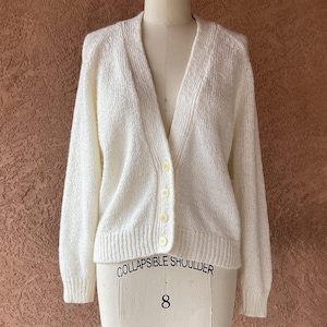May include: Cream-colored, long-sleeved cardigan sweater with a V-neck and button closure. The sweater is displayed on a dress form with the text "COLLAPSIBLE SHOULDER" and the number "8". The background is a textured, reddish-brown wall.
