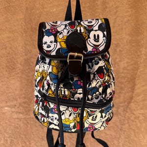 Puede incluir: Una pequeña mochila en blanco y negro con un patrón repetido de personajes de dibujos animados, incluyendo Mickey Mouse y el Pato Donald. La mochila tiene una solapa negra con cierre de hebilla y correas ajustables negras.