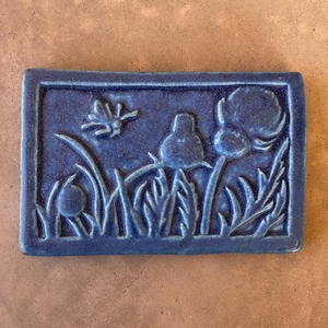 Könnte beinhalten: Eine rechteckige, kobaltblaue Keramikfliese mit einem erhabenen Reliefdesign. Das Design zeigt stilisierte Pflanzen, darunter Blumen und Gras, sowie ein kleines Insekt. Die Fliese ist von einem erhabenen Rand umgeben und liegt auf einer braunen Oberfläche.