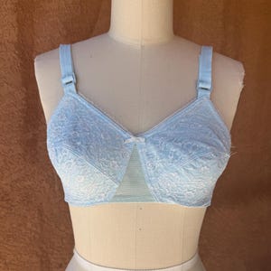 May include: A light blue bra featuring a floral lace overlay on the cups, a small center bow detail, and adjustable shoulder straps. The garment includes a solid mesh panel at the center gore and is displayed on a neutral-toned mannequin torso.