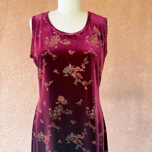 Peut inclure: Une robe sans manches en velours bordeaux profond ornée d'un motif floral répétitif dans des tons sourds de rose, d'or et de vert. Le vêtement présente une encolure arrondie et une silhouette droite en colonne descendant des épaules.