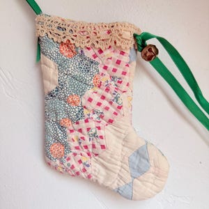 May include: A quilted Christmas stocking featuring a patchwork design of various floral and gingham patterns in shades of pink, blue, and orange. The top edge is trimmed with beige crochet lace and includes a green ribbon hanger with a small metal bell.