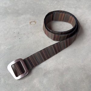 Puede incluir: Un cinturón de tela tejida con un patrón de rayas en color marrón oscuro y negro. El cinturón está enrollado e incluye una hebilla rectangular de metal plateado con esquinas redondeadas, diseñada para asegurar la correa a través del marco.