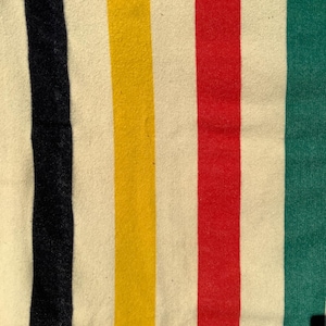 Puede incluir: Un tejido de color crema que presenta una serie de rayas verticales en negro, amarillo, rojo y verde. La tela tiene una textura suave y ligeramente difusa, y las rayas están espaciadas uniformemente sobre el fondo claro del material.
