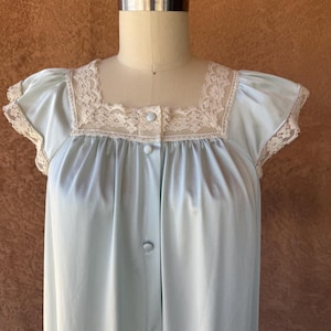 May include: A light blue nightgown featuring a square neckline and short cap sleeves trimmed with cream-colored lace. The garment has a front button placket with matching light blue buttons extending from the neckline down the center chest.
