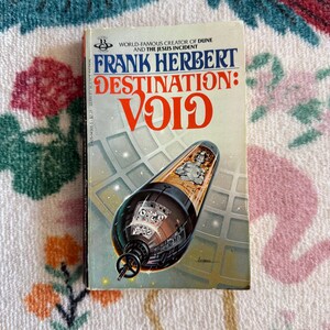Puede incluir: Un libro de bolsillo titulado Destination: Void de Frank Herbert. La portada muestra una nave espacial cilíndrica oscura flotando en el espacio sobre un fondo de cuadrícula, con una figura visible dentro de una sección transparente.