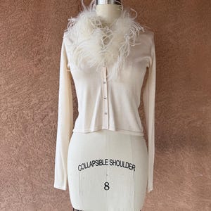 Pode incluir: Cardigã creme com mangas compridas e gola de penas brancas e fofas. O suéter com botões é exibido em um manequim com o texto "COLLAPSIBLE SHOULDER" e o número "8".