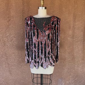 Pode incluir: Top vintage com lantejoulas com um padrão preto e vermelho. A blusa de manga comprida apresenta um decote redondo e é exibida em um manequim. As lantejoulas criam um design de listras verticais com uma bainha ondulada.