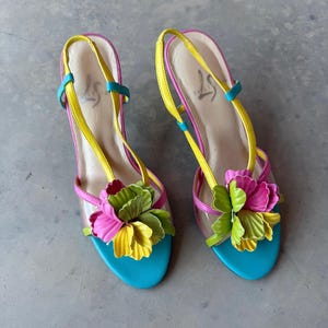 May include: A pair of slingback sandals featuring a bright turquoise sole, yellow straps, and a multi-colored floral accent on the front. The flower is composed of layered petals in shades of pink, lime green, and yellow. The insole is light beige.
