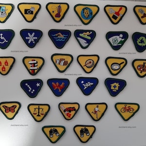 May include: A collection of 24 triangular Girl Guide badges with various embroidered designs, including a snowflake, a camera, a paintbrush, a recycling symbol, a butterfly, and a pair of binoculars. The badges are displayed on a white background with the text "JextIsland.etsy.com" repeated throughout.