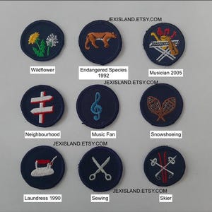 May include: A selection of navy blue embroidered patches, each featuring a different design. The patches depict a range of motifs, including flowers, a fox, musical instruments, a signpost, a treble clef, snowshoes, an iron, scissors, and skis. Text includes various descriptions.