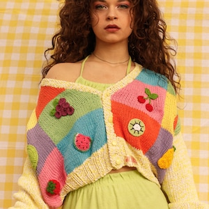 Op de afbeelding: Een vrouw draagt een kleurrijk gebreid vest met fruitapplicaties. Het vest is gemaakt van verschillende gekleurde vierkanten met een geel, wit en groen patroon. Het vest heeft een knoopsluiting en is gecropped.