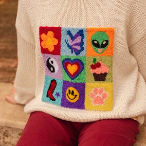 May include: A white knitted sweater with a colorful embroidered patch featuring various symbols, including a flower, butterfly, alien, yin yang, heart, cupcake, boot, smiley face, and paw print.