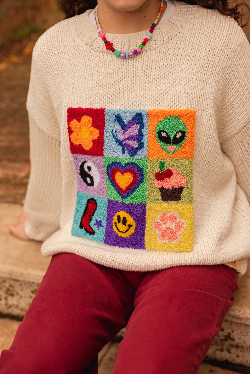 May include: A white knitted sweater with a colorful embroidered patch featuring various symbols, including a flower, butterfly, alien, yin yang, heart, cupcake, boot, smiley face, and paw print.