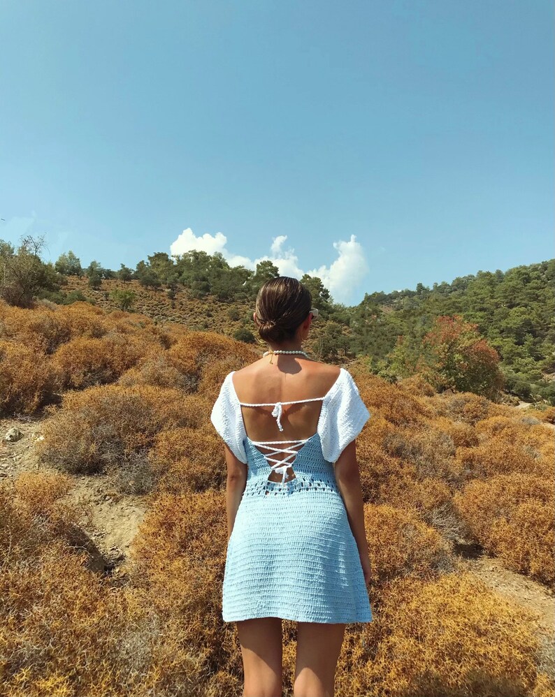 May include: A woman wearing a light blue crochet dress with white crochet detailing on the shoulders and a white crochet tie at the back. The dress has a lace-up back.