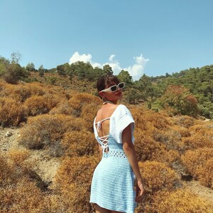 May include: A woman wearing a light blue crochet dress with white crochet detailing on the shoulders and a white lace-up back. She is wearing white sunglasses and is standing in a field of dry grass.