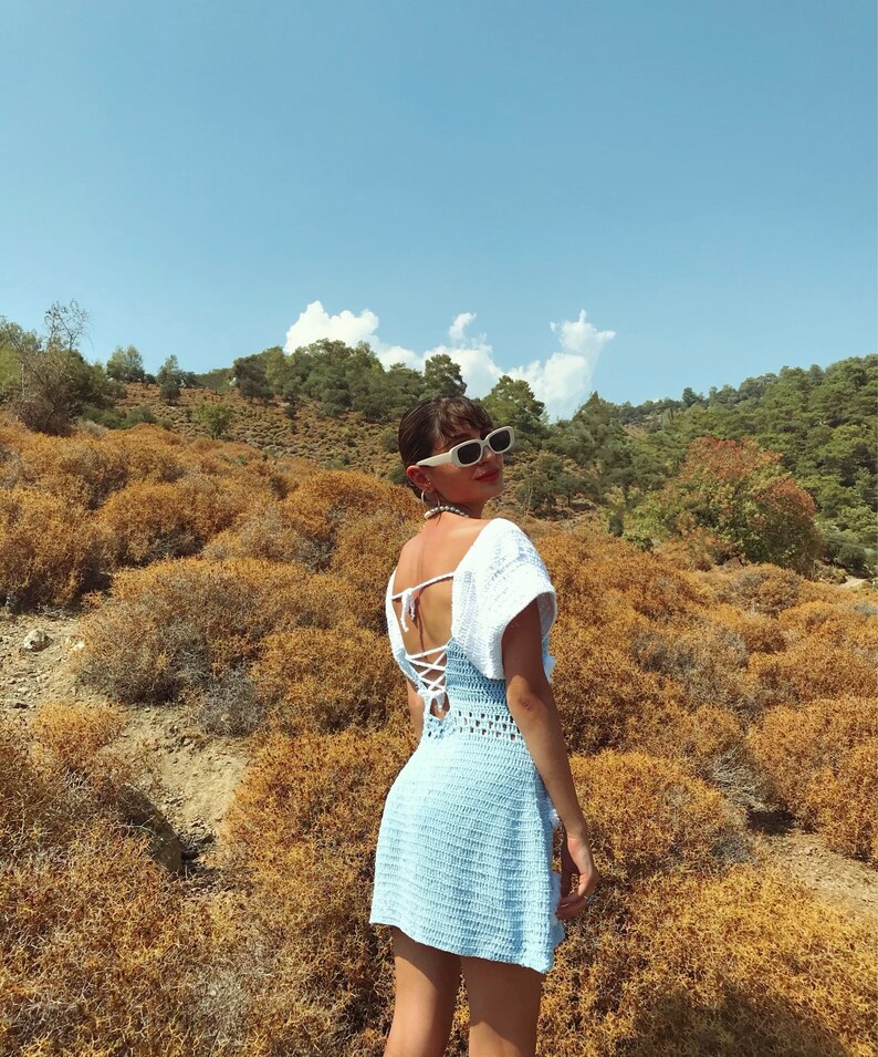 May include: A woman wearing a light blue crochet dress with white crochet detailing on the shoulders and a white lace-up back. She is wearing white sunglasses and is standing in a field of dry grass.