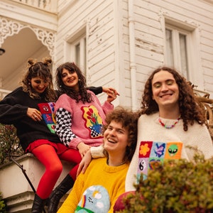 May include: Four friends wearing colorful sweaters with embroidered designs stand in front of a white building. The sweaters feature various patterns, including flowers, rainbows, and geometric shapes.