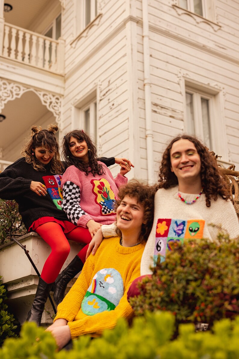May include: Four friends wearing colorful sweaters with embroidered designs stand in front of a white building. The sweaters feature various patterns, including flowers, rainbows, and geometric shapes.