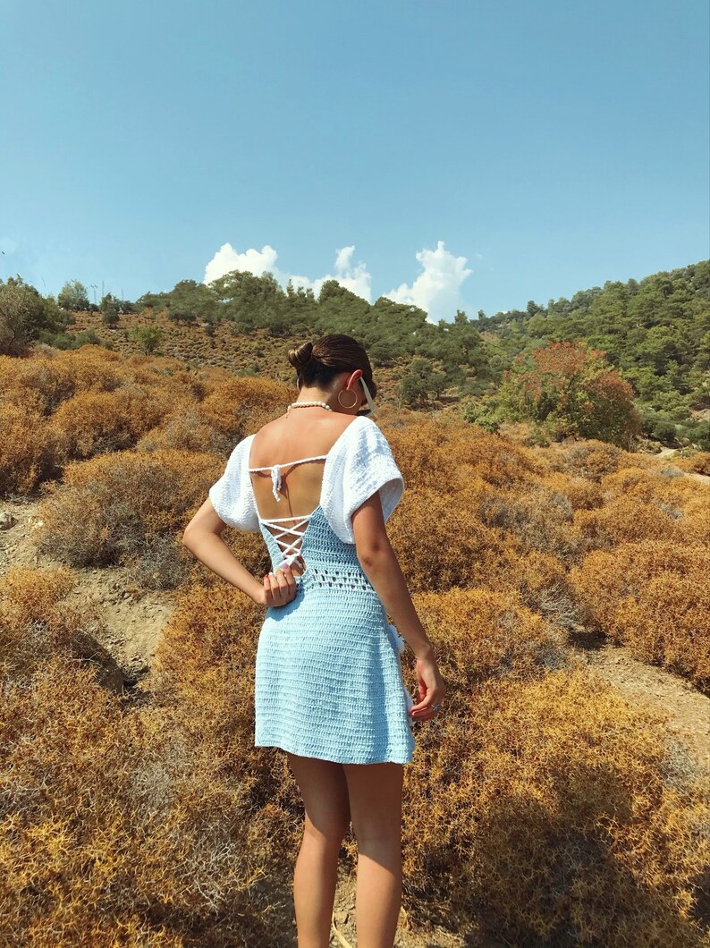 May include: A woman wearing a light blue crochet dress with a white crochet top and a white ribbon tied at the back. She is standing in a field of dry grass with a blue sky and white clouds in the background.