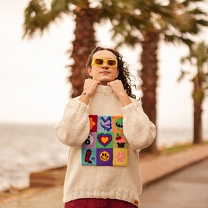May include: A person wearing a white sweater with a colorful patch of embroidered designs, including a heart, a butterfly, a cupcake, and a smiley face. They are wearing red pants and yellow sunglasses.