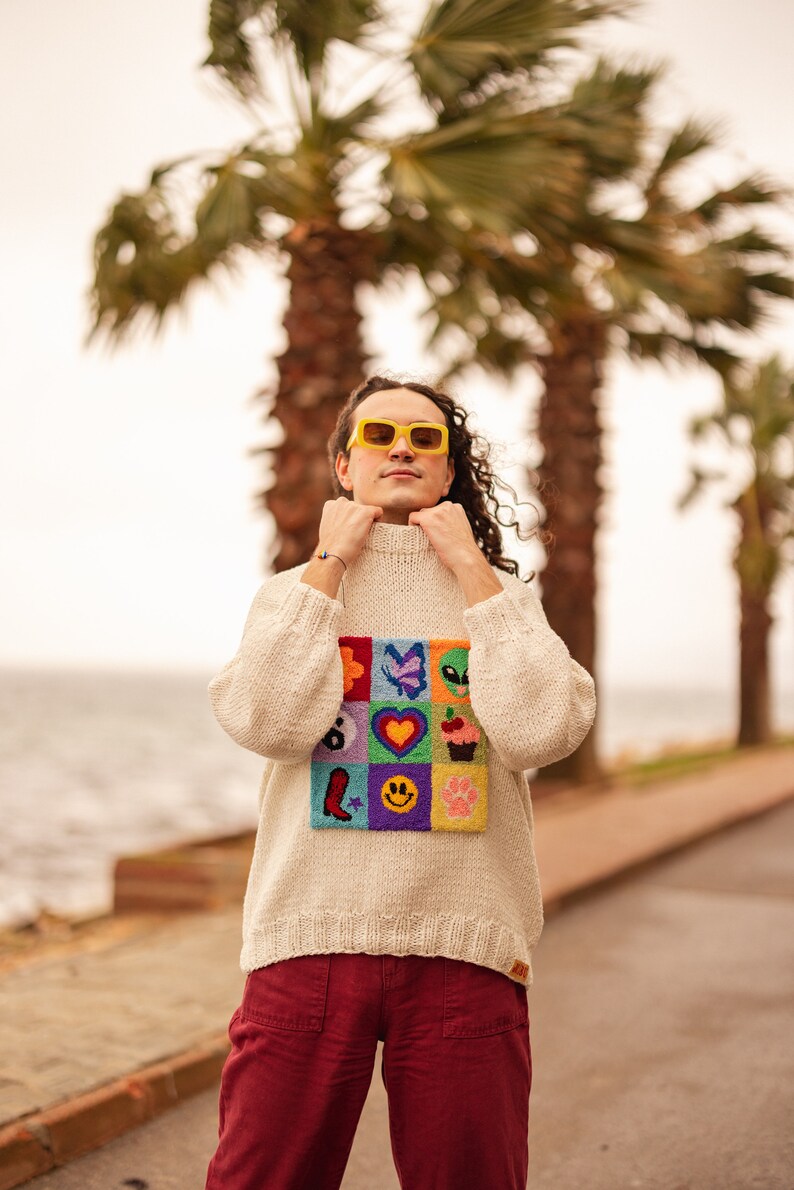 May include: A person wearing a white sweater with a colorful patch of embroidered designs, including a heart, a butterfly, a cupcake, and a smiley face. They are wearing red pants and yellow sunglasses.