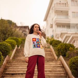 May include: A person wearing a white sweater with a colorful patch design and red pants stands on a set of stairs. The sweater features a variety of colorful patches, including a flower, a butterfly, a yin-yang symbol, a heart, and a smiley face.