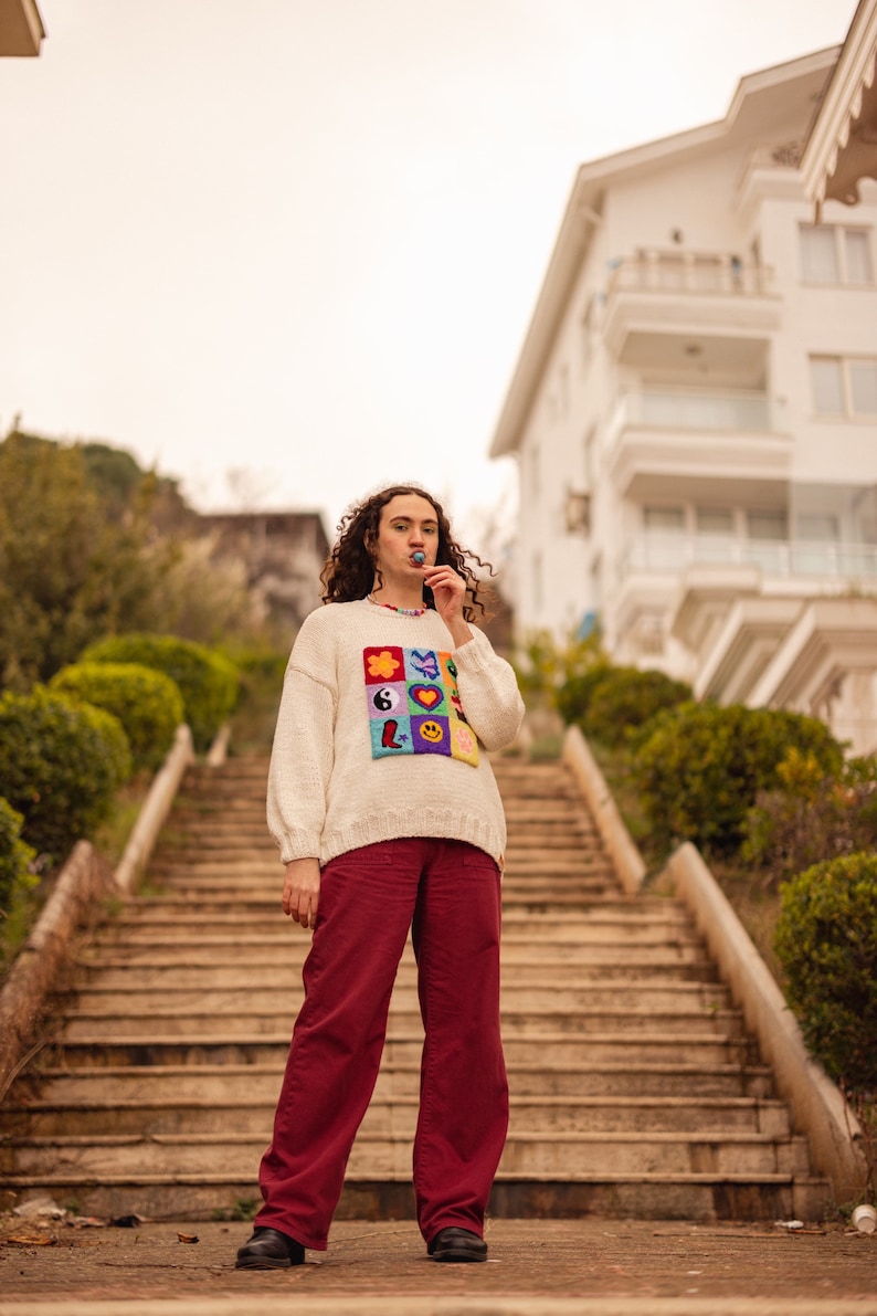 May include: A person wearing a white sweater with a colorful patch design and red pants stands on a set of stairs. The sweater features a variety of colorful patches, including a flower, a butterfly, a yin-yang symbol, a heart, and a smiley face.