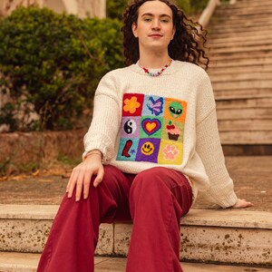 May include: A person wearing a white sweater with a colorful embroidered patch of various symbols, including a flower, butterfly, alien, yin yang, heart, cupcake, star, smiley face, and paw print. They are wearing red pants and black shoes and sitting on a set of stone steps.