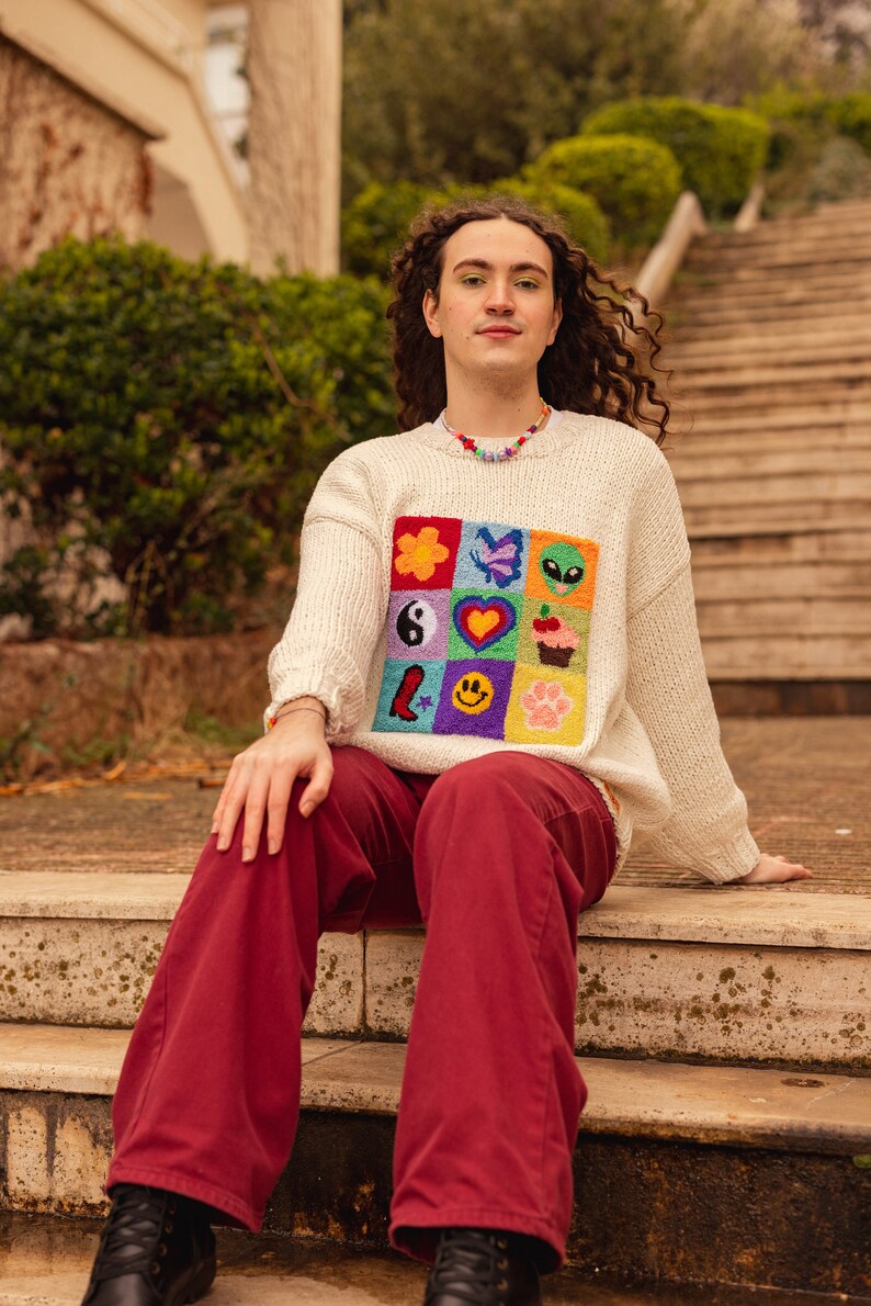 May include: A person wearing a white sweater with a colorful embroidered patch of various symbols, including a flower, butterfly, alien, yin yang, heart, cupcake, star, smiley face, and paw print. They are wearing red pants and black shoes and sitting on a set of stone steps.