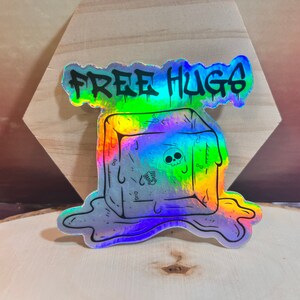 Puede incluir: Una pegatina holográfica con un cubo de hielo derritiéndose y el texto "FREE HUGS" en negro.