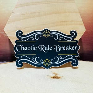 May include: A black sticker with the text "Chaotic Rule Breaker" in white script. The sticker has a decorative border with white swirls and yellow geometric shapes. The sticker is on a wooden surface.