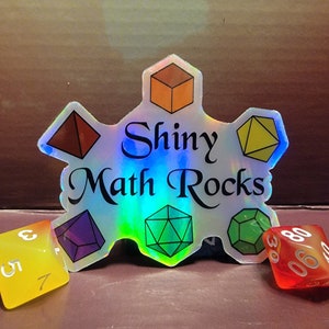 Puede incluir: Una pegatina holográfica con el texto "Shiny Math Rocks" rodeada de formas geométricas de colores. Hay dos dados en primer plano, uno amarillo y otro rojo.