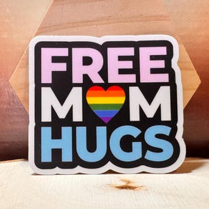 以下が含まれることがあります： 太字のブロックフォントで「FREE MOM HUGS」という言葉が書かれたステッカー。 「FREE」は薄いピンク色、「MOM」は白、「HUGS」は水色です。 「MOM」という言葉の中央には虹色のハートがあります。
