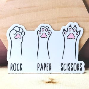 May include: A white sticker featuring three cat paws in the classic Rock, Paper, Scissors game poses. The paws have pink paw pads and are outlined in black. The words "ROCK", "PAPER", and "SCISSORS" are printed below each paw.