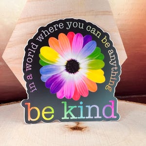 以下が含まれることがあります： 虹色の花に「in a world where you can be anything be kind」という文字が書かれています。花は黒い背景の上にあります。