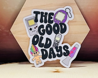 May include: A sticker with the text "The Good Old Days" in black with a retro 90s theme. The sticker features a purple Tamagotchi, a Game Boy, a troll doll, a cell phone, and a lava lamp.