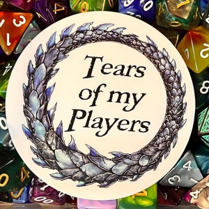 Puede incluir: Una pegatina blanca con un diseño de cola de dragón azul y el texto "Tears of my Players". La pegatina está rodeada de dados de colores.