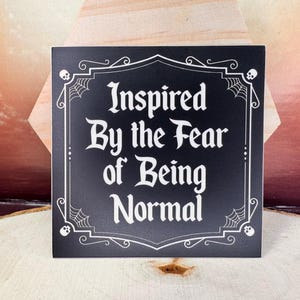 May include: Black and white sign with a gothic-style border and the text "Inspired By the Fear of Being Normal".