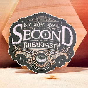 May include: A black sticker with the text "But what about Second Breakfast?" in white, stylized lettering. The sticker has an ornate design with a ring illustration and is set against a wooden background.