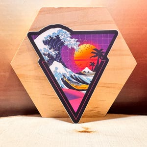 May include: A sticker with a retro-style design featuring a large ocean wave, a setting sun, and palm trees. The image is set within a black-outlined triangle with a purple grid background. The sticker is placed on a wooden surface.