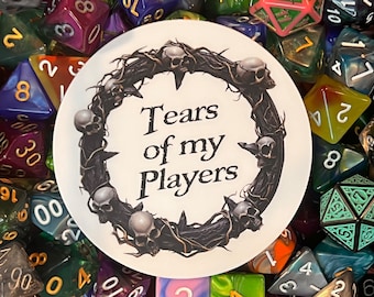May include: A white round sticker with a black wreath of thorns and skulls around the text "Tears of my Players". The sticker is surrounded by colorful dice.