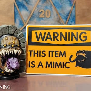 May include: A yellow warning sign with black text that reads "WARNING THIS ITEM IS A MIMIC". A black silhouette of a monster with a large mouth and teeth is on the right side of the sign. A small, gray, stone-like figure with a large mouth and purple tongue is in the background.