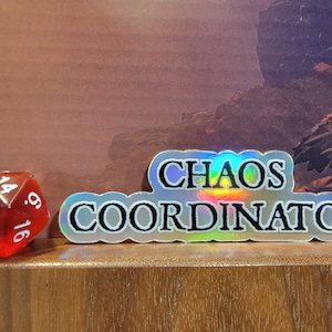 Op de afbeelding: Een holografische sticker met de tekst "Chaos Coordinator" in zwarte letters op een zilveren achtergrond. Op de voorgrond ligt een rode twintigzijdige dobbelsteen.